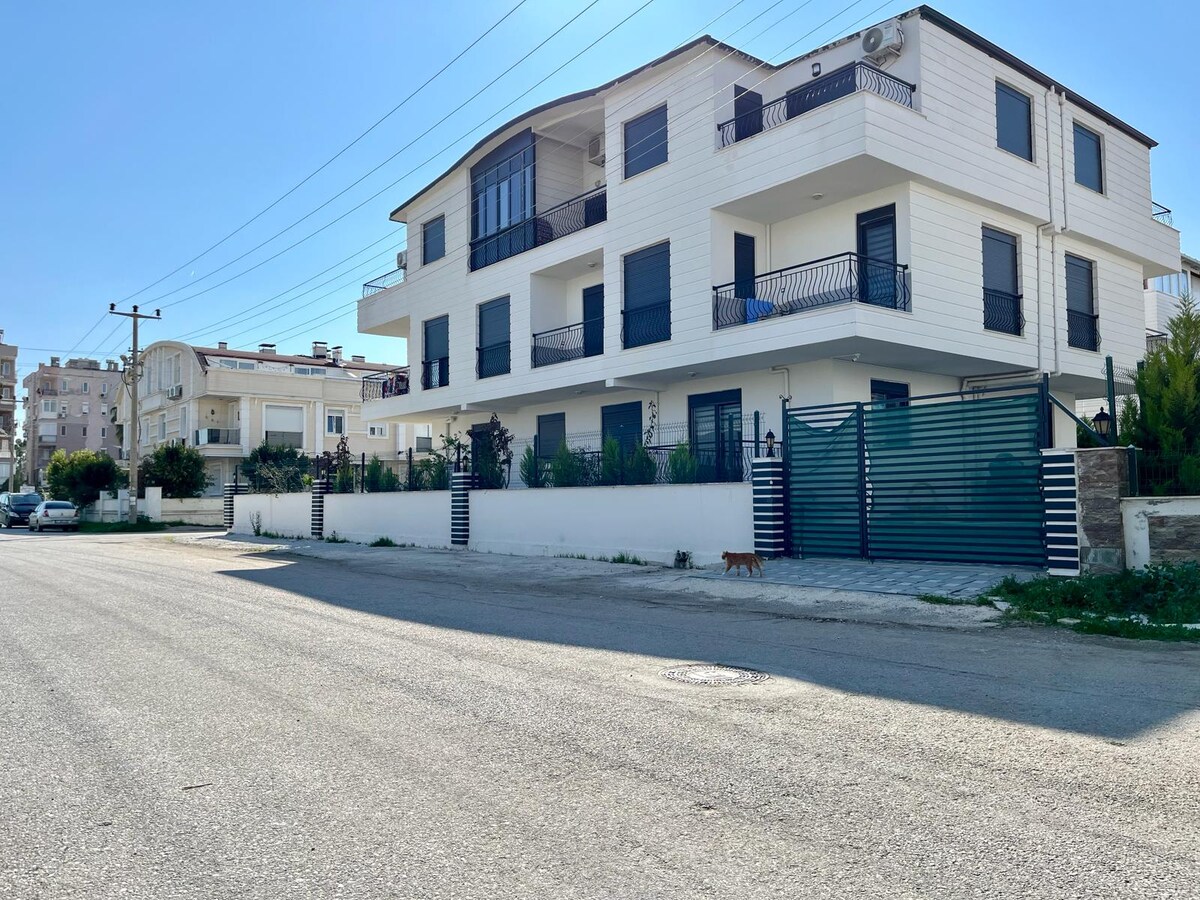 The exterior of the newly built flat is presented, showcasing a three-story white structure with multiple balconies. A gated entrance is visible, along with a landscaped area. The surrounding street is quiet, with neighboring buildings and vehicles parked along the curb.