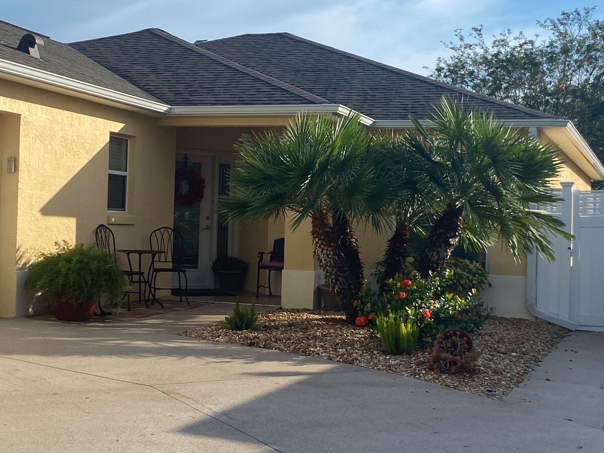 The exterior of the villa features a welcoming entrance with a small patio. Two black chairs are positioned by the door, surrounded by lush palm trees and colorful plants. Gravel landscaping is visible, enhancing the home's curb appeal.