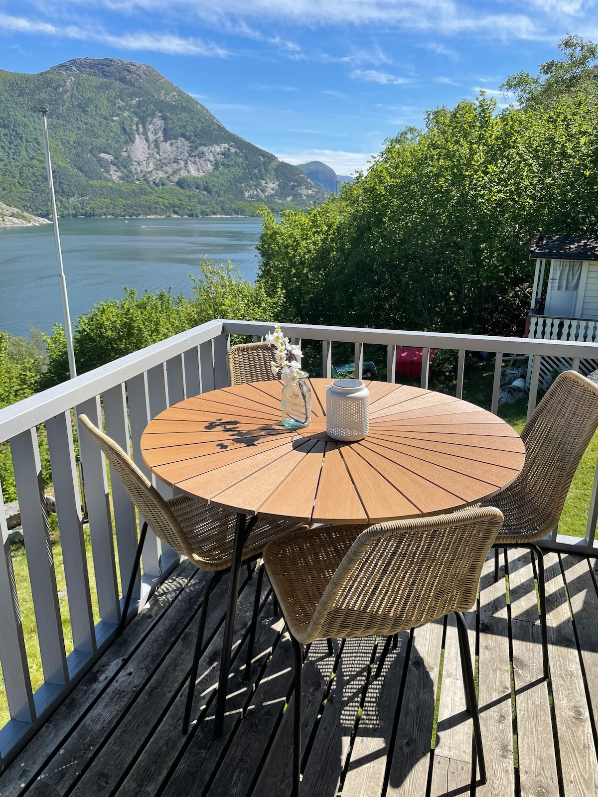 A round wooden table is set on a deck, surrounded by four woven chairs. A vase with flowers and a lantern are centered on the table. Scenic views of the mountains and water are visible in the background, enhancing the outdoor dining space.