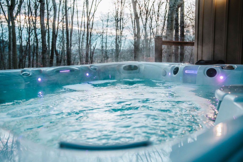 A hot tub is situated on a porch overlooking a serene forested landscape. Soft ambient lighting lines the interior wall of the tub, reflecting off the clear water. The surrounding trees create a peaceful environment, inviting relaxation.