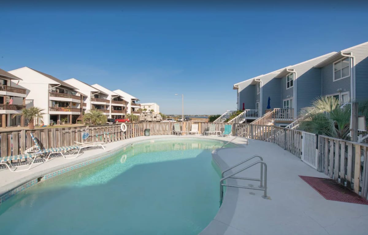 A semi-private pool is surrounded by a wooden fence, with lounge chairs positioned along the patio. Nearby townhome structures feature multiple levels, and palm trees add a touch of greenery. The calm water reflects the clear blue sky and creates a relaxing environment.