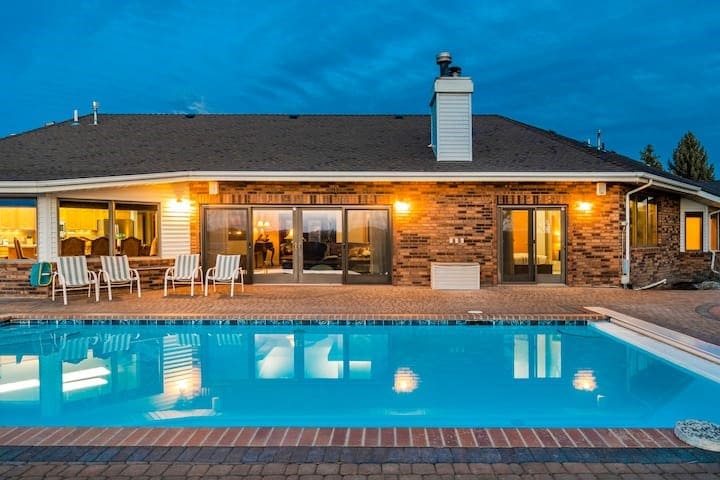 The exterior of the property is presented with a heated pool in the foreground, reflecting the evening sky. A spacious patio area features several lounge chairs. The house is illuminated by warm lights, and large glass doors invite views into the interior space.