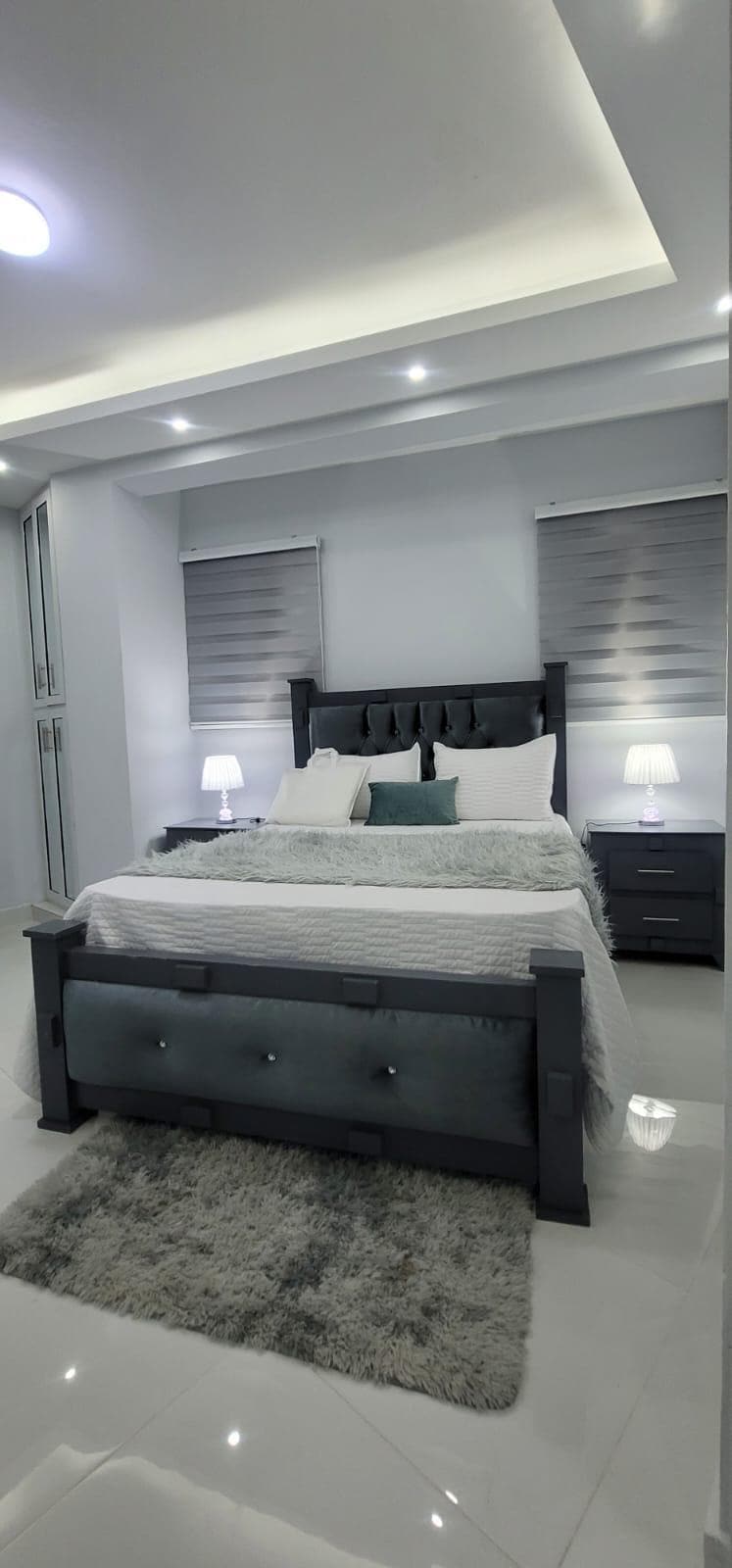 A well-appointed bedroom features a spacious bed with light linens and decorative pillows. Twin bedside lamps provide soft illumination. The sleek furnishings include side tables, and stylish grey curtains complement the modern aesthetic. A plush rug lies beneath the bed, enhancing the comfortable ambiance.