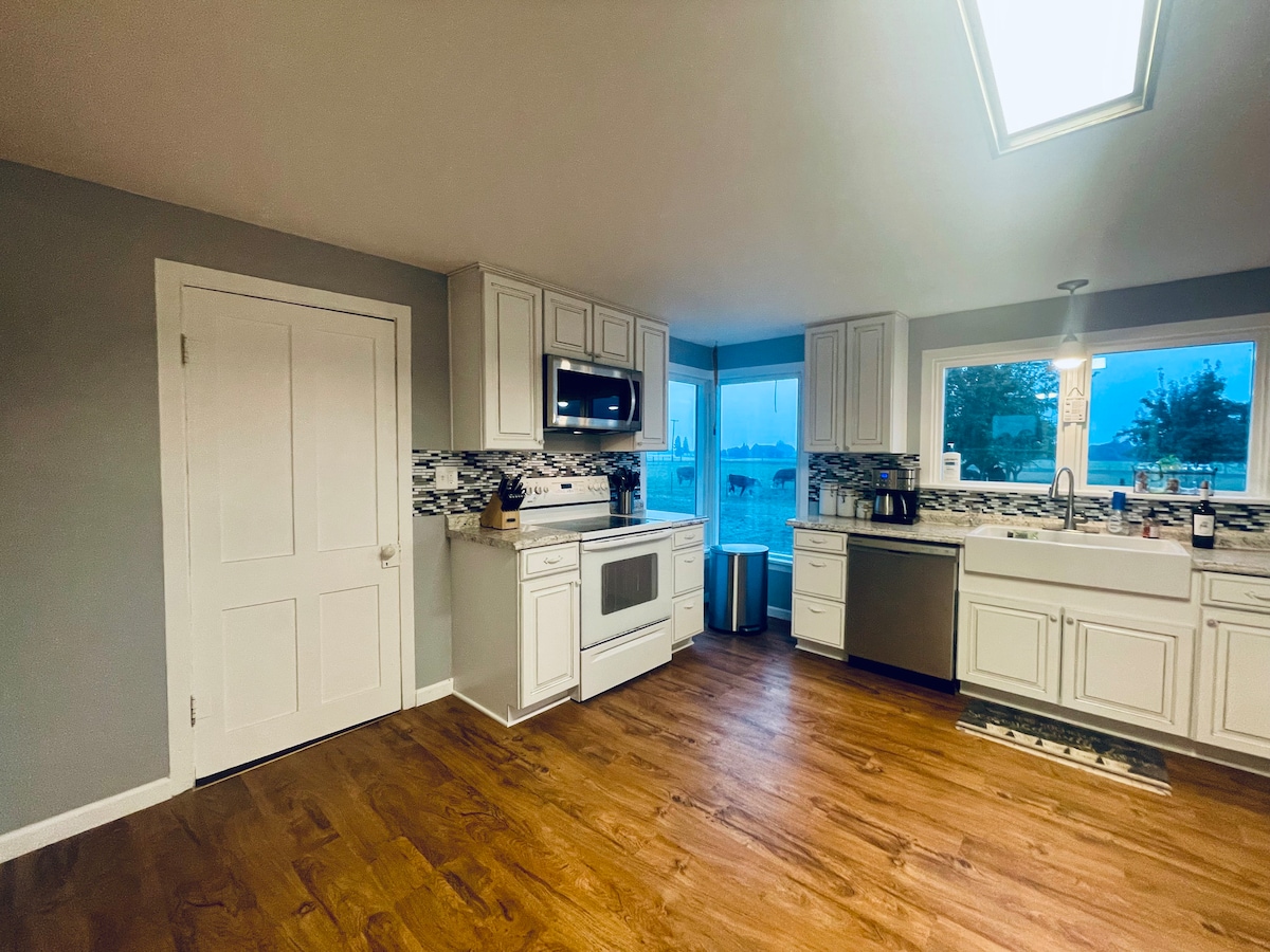 The kitchen features white cabinetry with a modern backsplash and stainless-steel appliances. A window allows natural light to enter and provides views of the outdoors. The flooring is a warm wood tone, enhancing the spacious feel of the area.