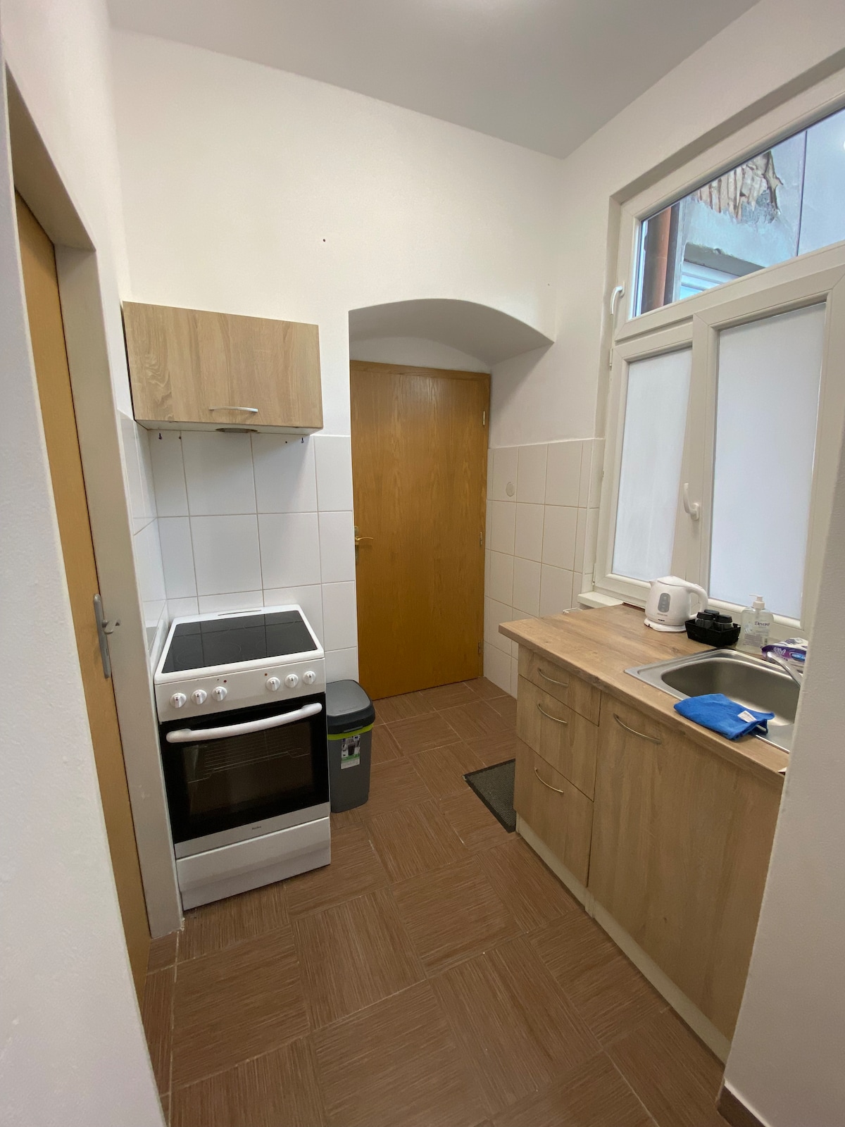 A compact kitchen area is shown, featuring a wooden cabinet above the stove and a countertop with a sink. An oven and stove are present alongside a microwave and kettle. Large windows provide natural light, while tiled walls enhance the clean, simple design.