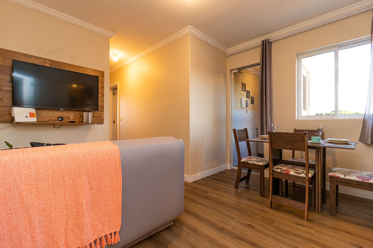 An inviting living space features a comfortably arranged seating area with a soft gray sofa, complemented by an orange throw. A dining table for four sits nearby, with floral-patterned chairs. A wall-mounted television adds to the entertainment options, with ample natural light streaming through the window.