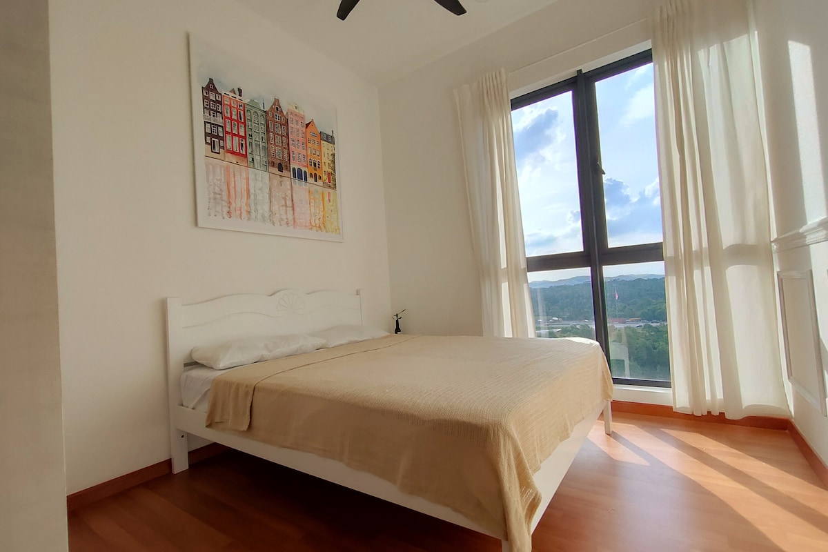 A well-lit bedroom features a queen-sized bed with a neutral-colored blanket. Large windows allow natural light to fill the space, and colorful artwork hangs above the bed. The room is designed with a light color palette and wooden flooring.