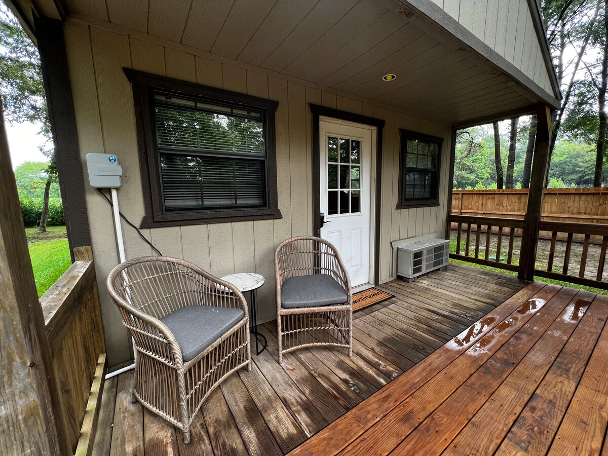 A covered porch area is shown with two wicker chairs positioned on a wooden deck. A front door with glass panels leads into the cottage. Nearby, an air conditioning unit sits mounted on the wall, and greenery is visible beyond the porch railing.