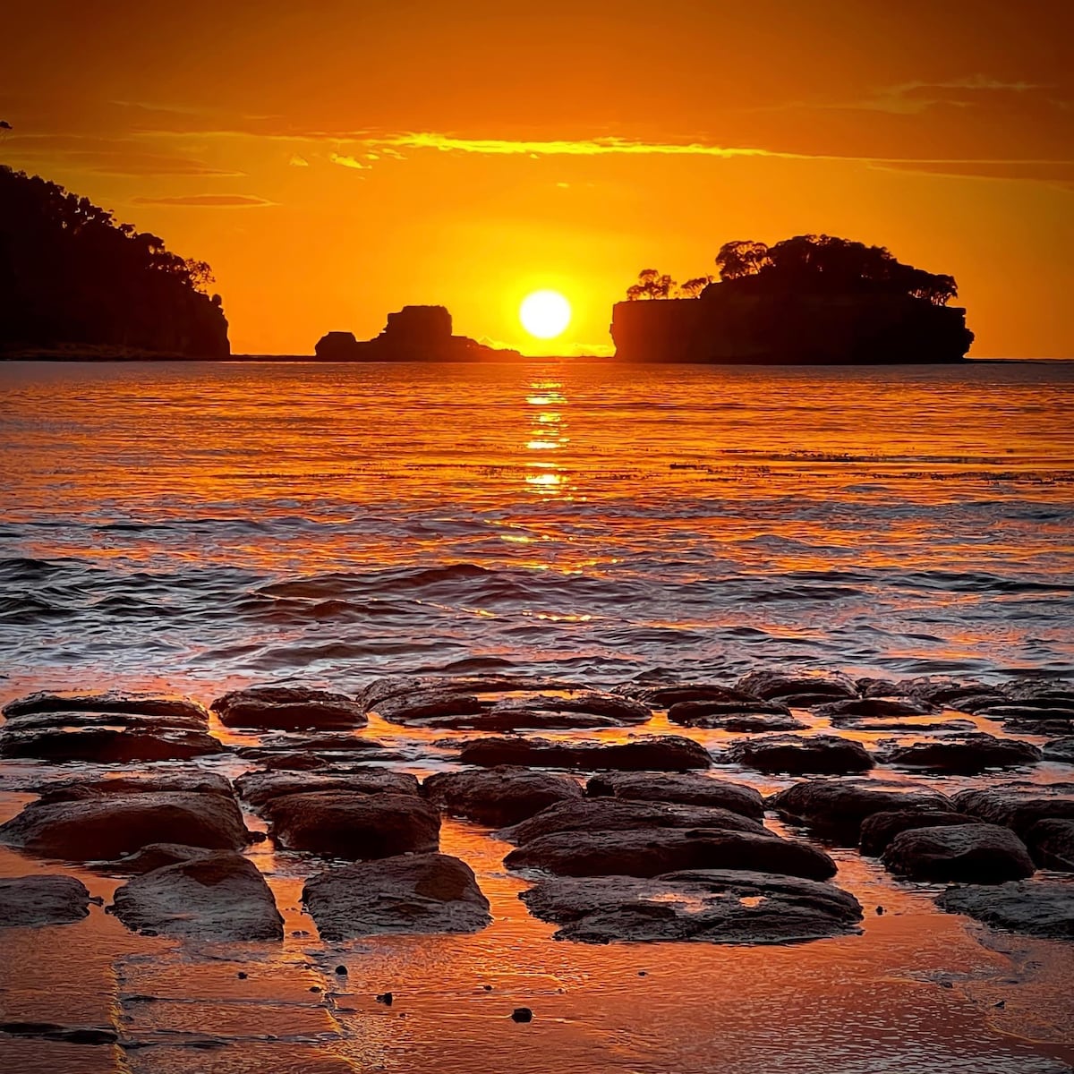 A stunning sunset casts warm orange and golden hues over the sea, reflecting on the water's surface. Silhouetted rock formations are visible in the distance against the colorful sky, while textured rocks in the foreground are glistening with moisture.