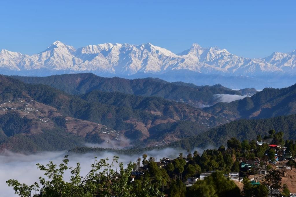 The image captures a stunning panorama of snow-capped mountains against a clear blue sky, with layers of green hills extending below. Soft clouds drift among the valleys, enhancing the serene landscape that stretches into the distance.