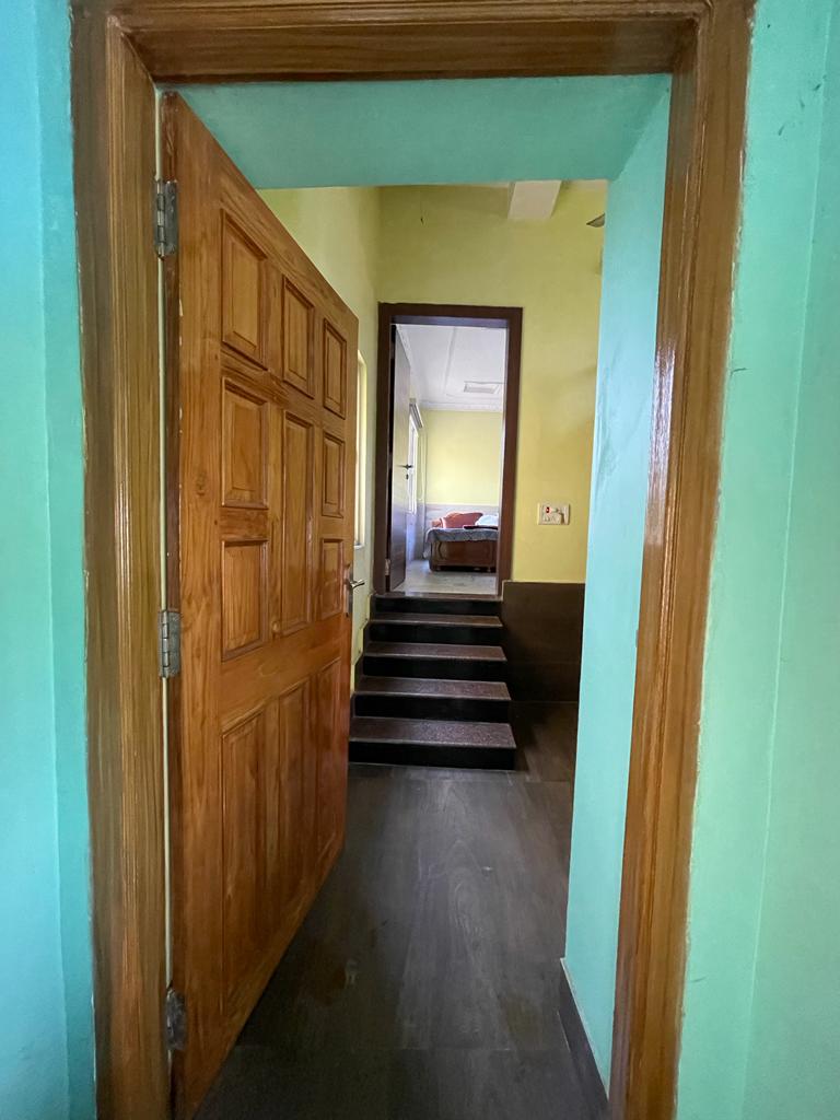 A wooden door is visible, leading to a hallway with painted green walls. Stairs ascend in the background, guiding toward a softly lit area with an undefined space beyond. The floor is dark, presenting a contemporary design.