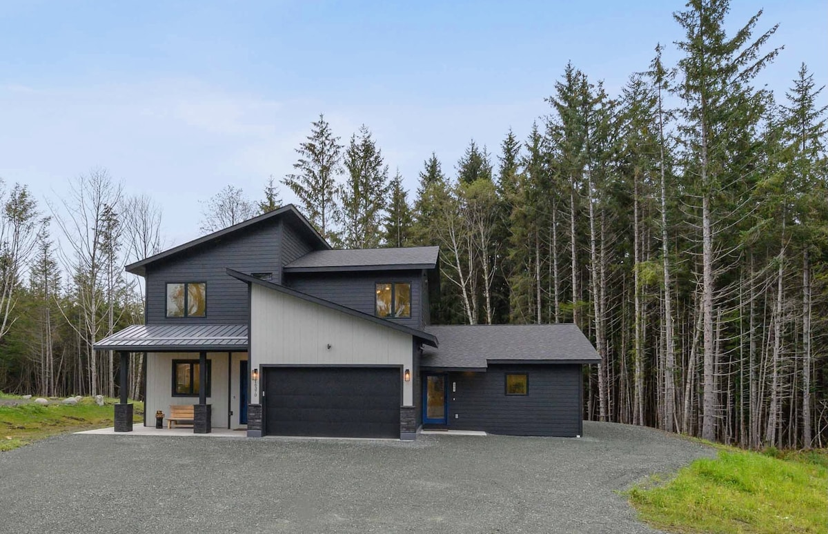 A modern two-story apartment building features a sleek black and gray exterior, surrounded by lush trees. A spacious gravel driveway leads to the entrance, which is accentuated by large windows and a stylish front door. The setting offers a tranquil woodland atmosphere.