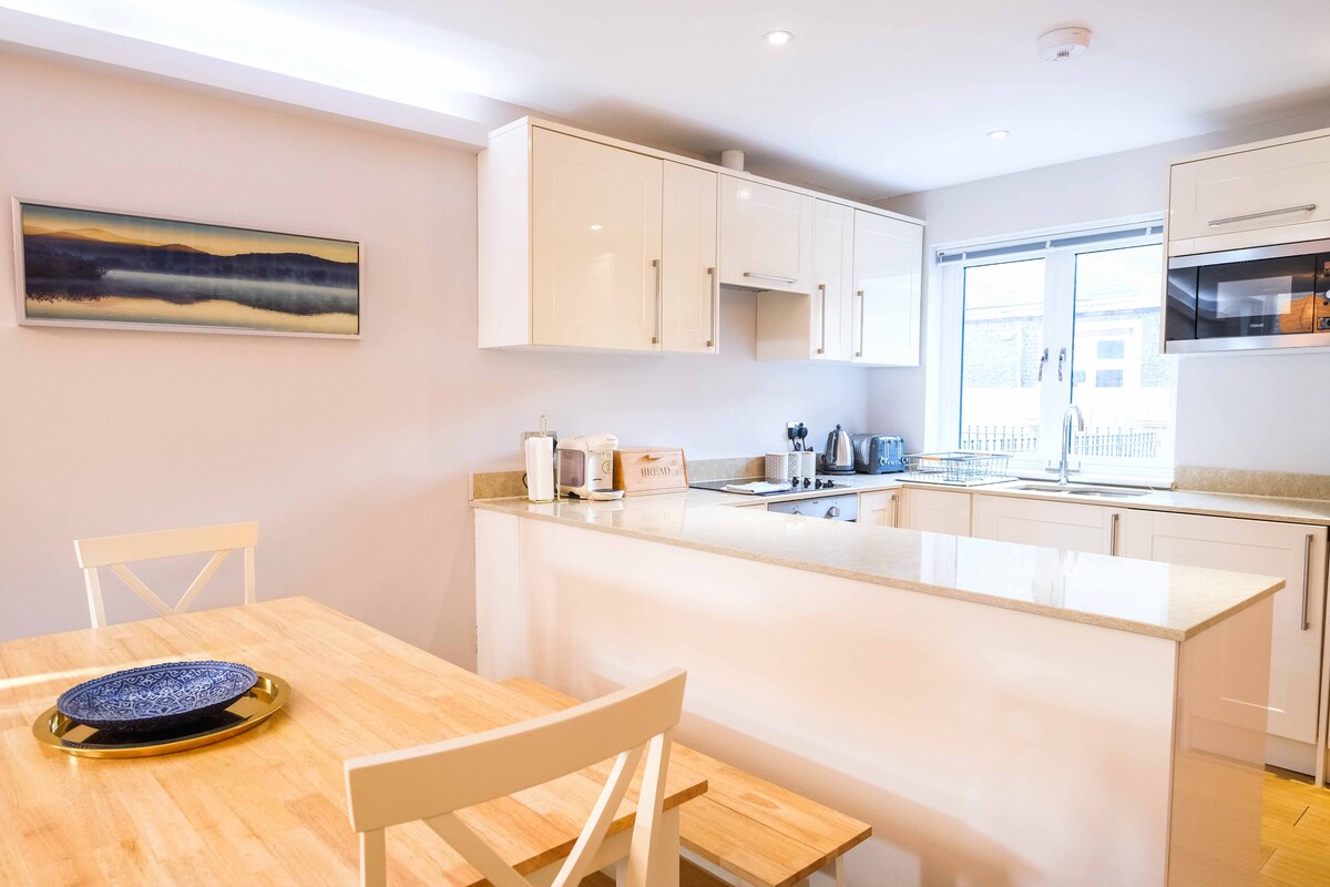 An open plan kitchen and living area is featured, with modern white cabinetry and a light countertop. A wooden table with two chairs is visible, accompanied by a blue plate. Natural light streams in from the window, illuminating the space.