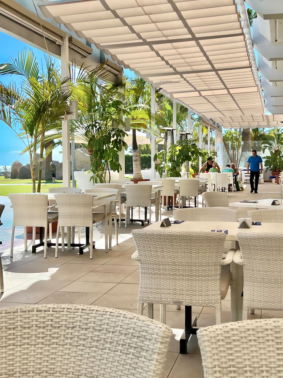 An airy dining area features white wicker tables and chairs, arranged under a sun-shaded canopy. Lush green plants are positioned throughout, and large windows allow natural light to fill the space. A waiter is visible attending to guests in the background.