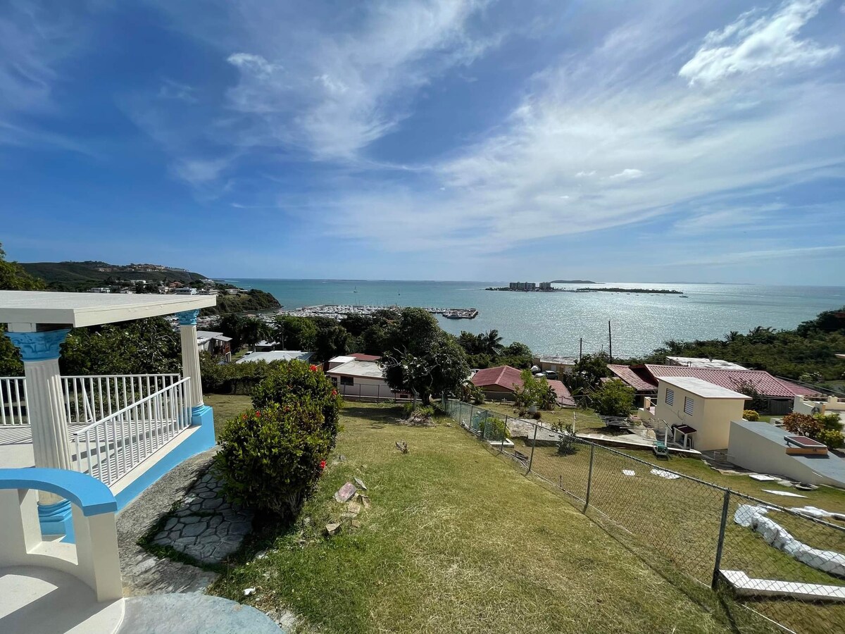 An expansive view overlooks the water where the Atlantic and Caribbean Seas converge. A grassy area with scattered plants leads to a porch with white and blue accents. In the distance, a marina is visible along the shoreline under a clear blue sky.