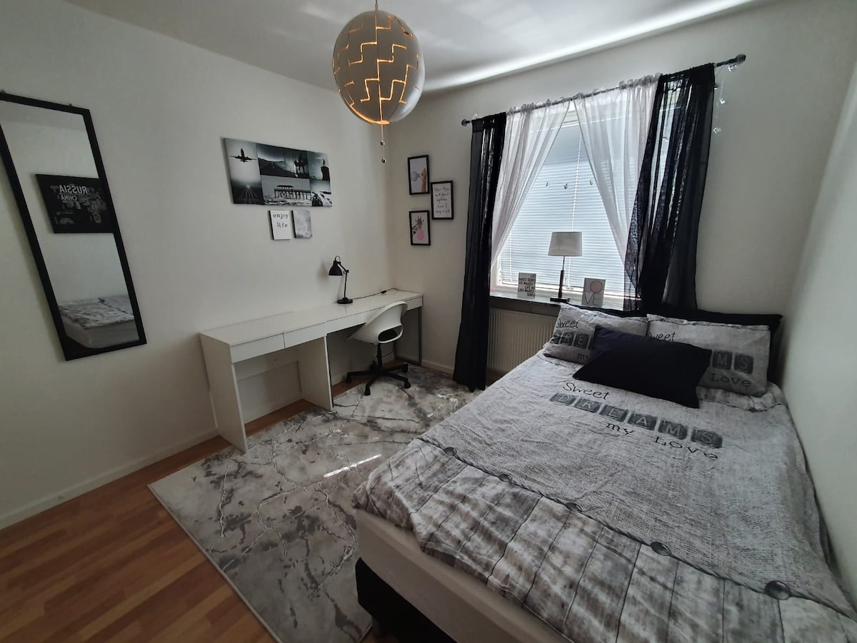 The well-lit bedroom features a spacious bed adorned with a patterned blanket. A desk with a chair is positioned near a window, while decorative wall art and a round pendant light add style. A large mirror reflects the room’s open layout.