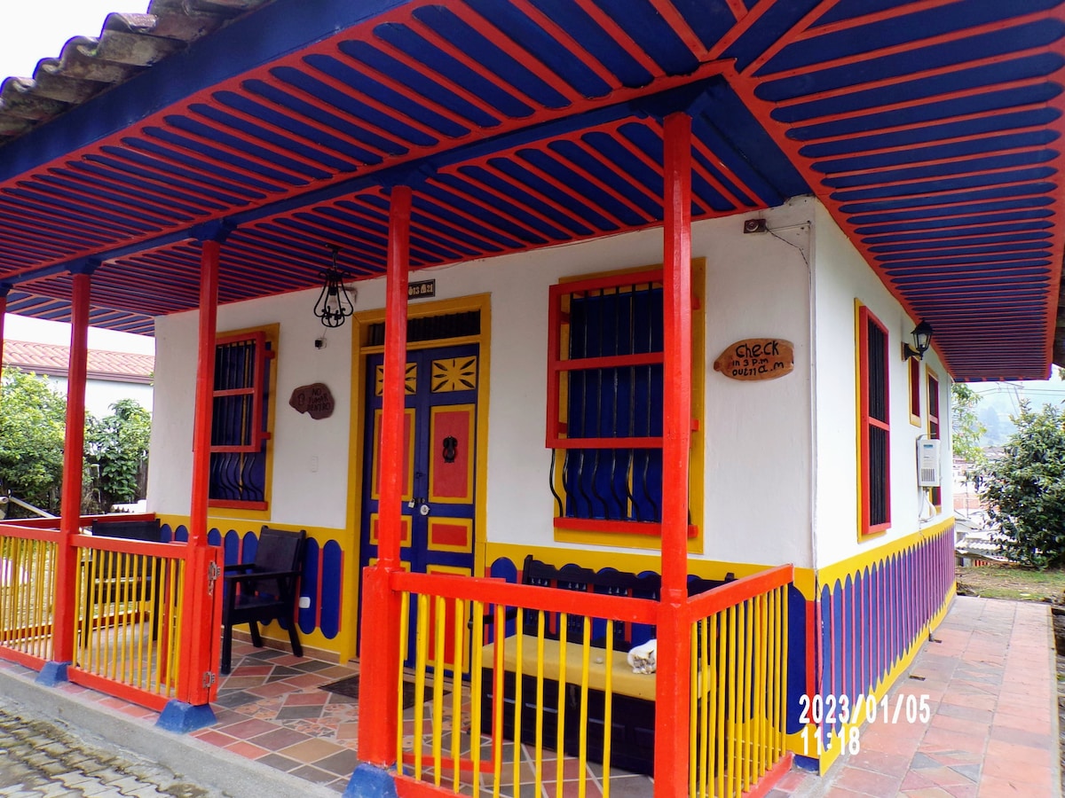 A vibrant cabin exterior is presented with colorful blue and yellow accents. Decorative elements include a welcoming door and multiple windows, which are adorned with classic shutters. A porch space with seating provides an inviting entry point surrounded by lush greenery.