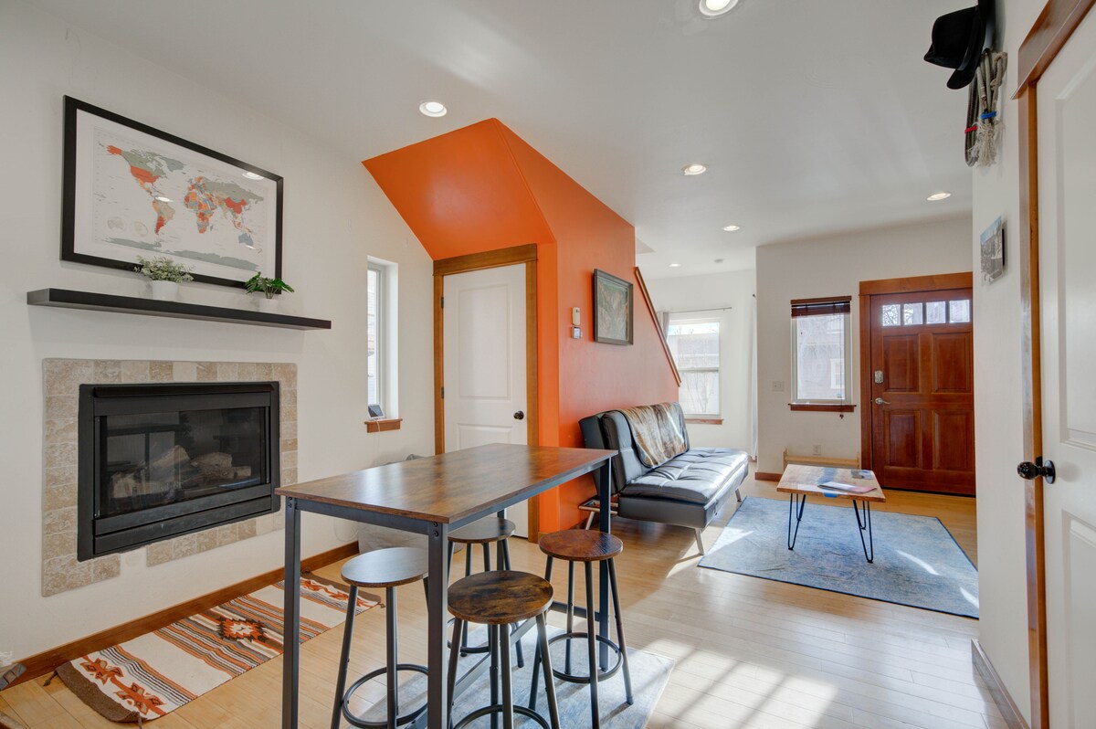 A bright living space is highlighted by an orange accent wall, complemented by a map artwork above the fireplace. A modern sofa is positioned near a coffee table, and a dining table with stools occupies the center of the area. Natural light filters through the windows.