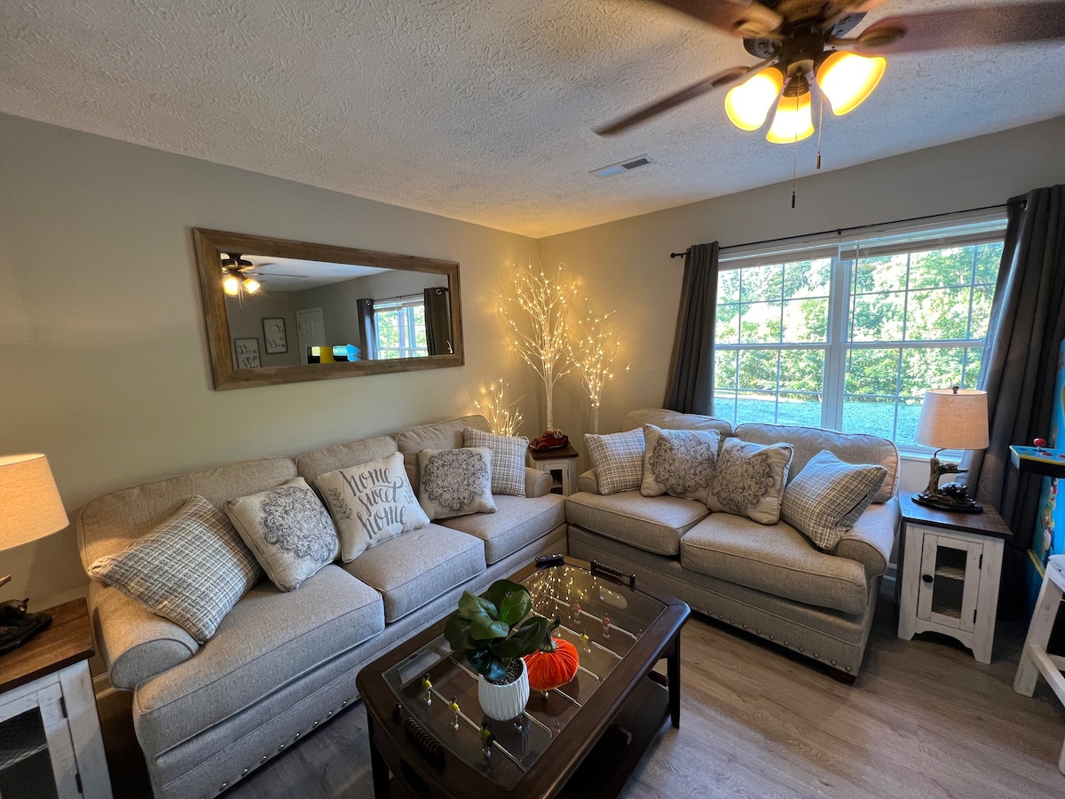 A spacious living area features a light grey sectional sofa adorned with decorative pillows. A wooden coffee table sits in front, complemented by a potted plant and seasonal decor. Soft lighting is provided by bedside lamps, enhancing the cozy atmosphere near a large window framed by dark curtains.
