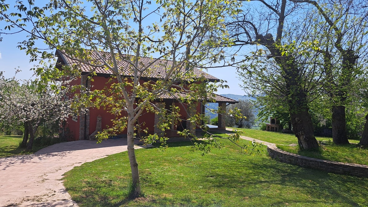 The exterior of the villa is highlighted by a red facade and a tiled roof. Surrounding greenery includes trees and well-maintained grass, enhancing the natural environment. A stone pathway leads to the entrance, with distant hills visible in the background.