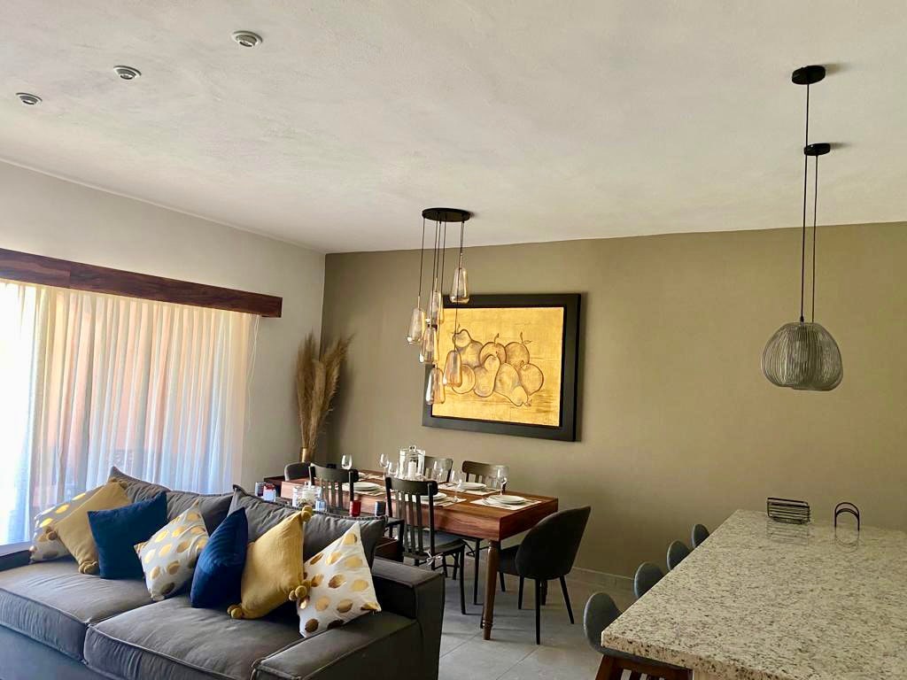 An open living area features a comfortable sofa adorned with decorative cushions in various shades. A dining table is set with tableware, complemented by a wall art piece that adds character. Large windows allow natural light to brighten the space.