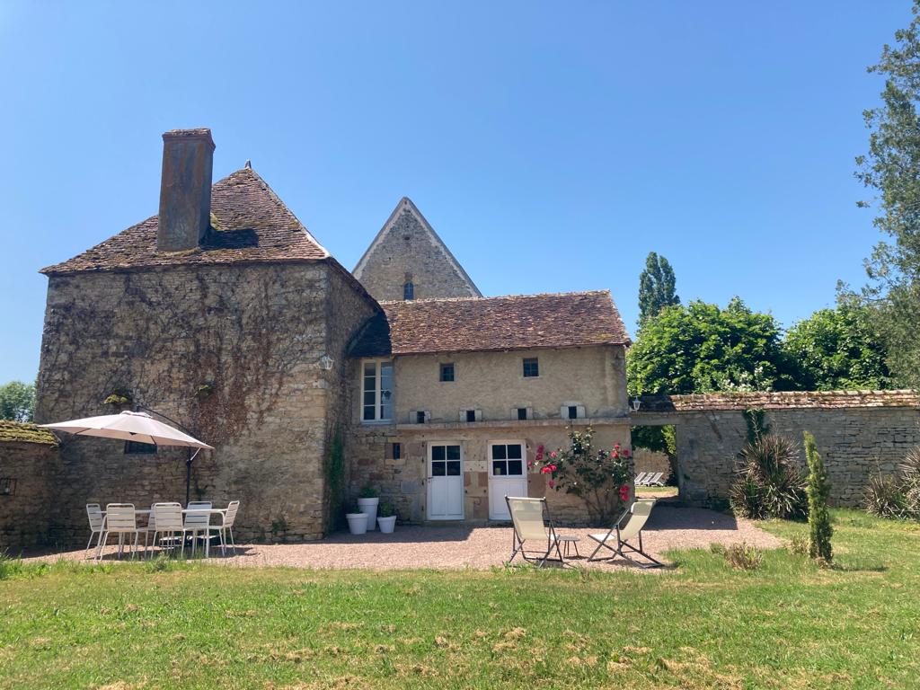 The exterior of the property showcases a charming stone building with a sloped roof. A gravel area features a dining table with chairs and lounge chairs, surrounded by greenery and flowers. The sun-filled space is ideal for outdoor relaxation.