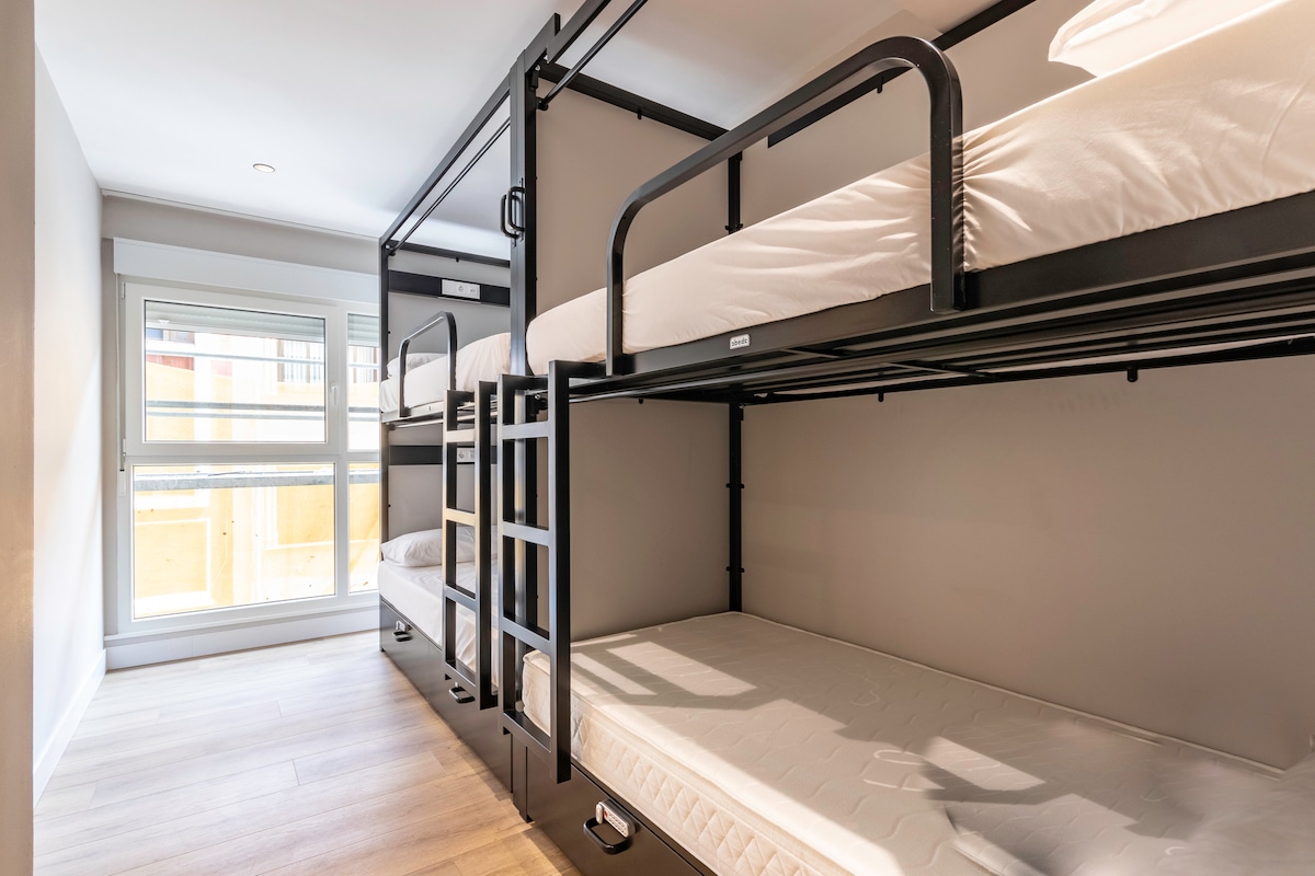 A shared room features two sets of black metal bunk beds with comfortable mattresses. Natural light fills the space through a large window, offering views of the outside. The room is designed with a neutral color palette, contributing to a clean and welcoming environment.