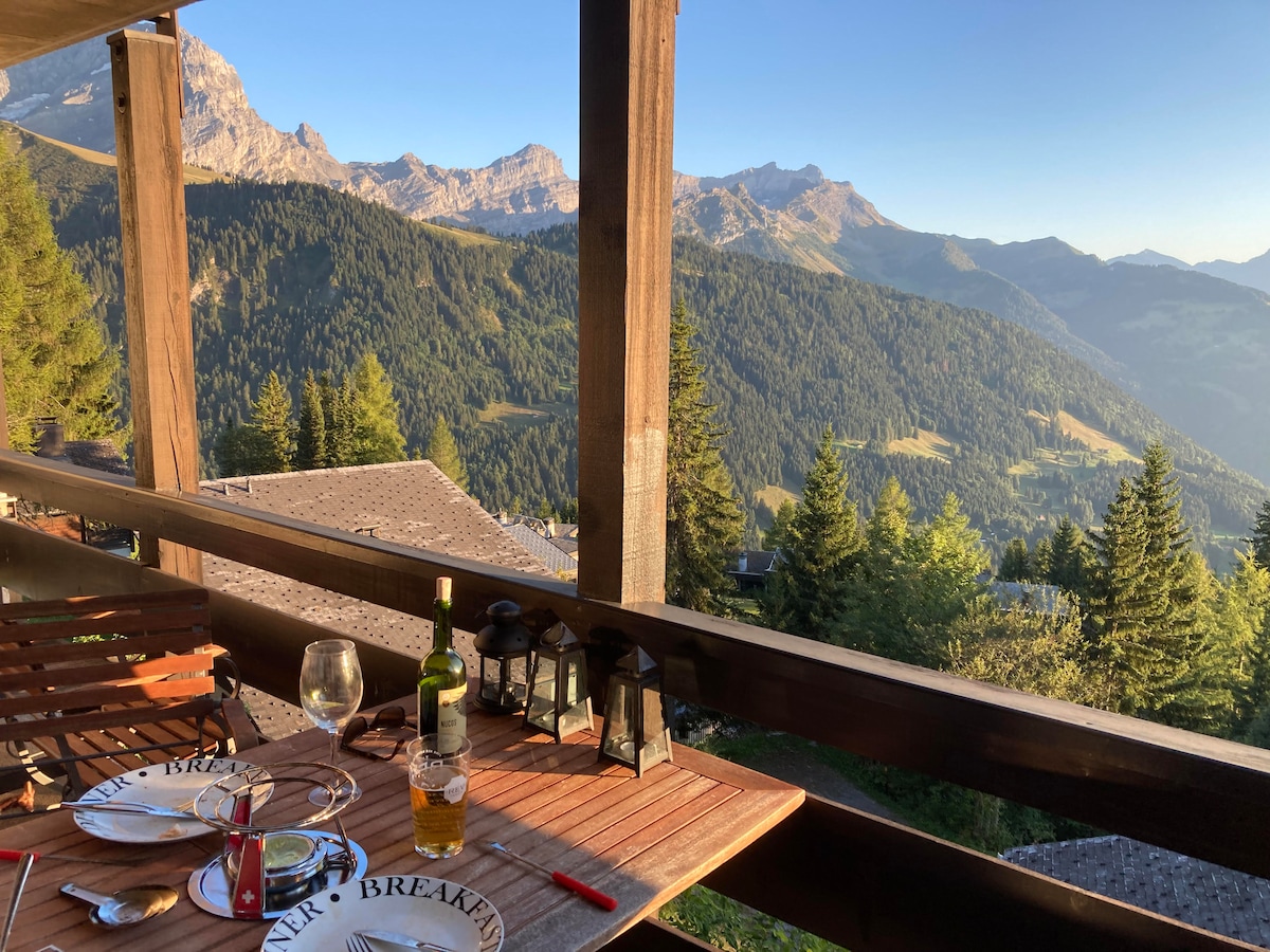 A spacious balcony is presented with a wooden table set for a meal, featuring plates, glasses, and bottles. Surrounding the area are lush trees and expansive views of the Swiss mountains, capturing the serene landscape under a clear sky.