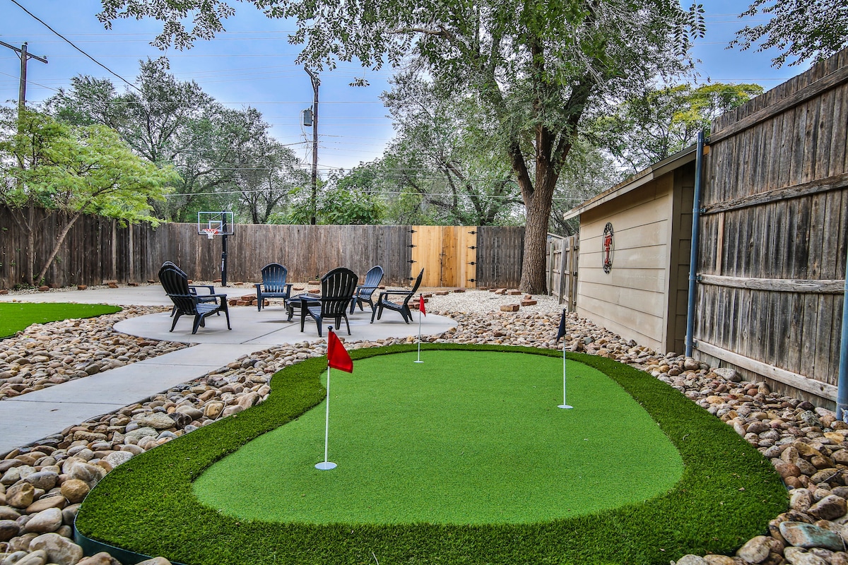 A backyard area is shown, featuring a well-maintained putting green with two flags and a seating arrangement of several black chairs surrounding a stone-paved space. A mini basketball hoop is visible in the background, surrounded by wooden fencing and abundant greenery.