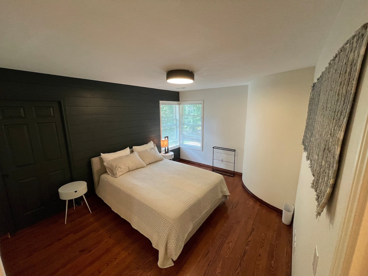 A spacious bedroom features a queen bed with light-coloured linens. A nightstand with a lamp is positioned beside the bed. Natural light enters through the large window, brightening the room, which is accented by dark walls and warm wood flooring.
