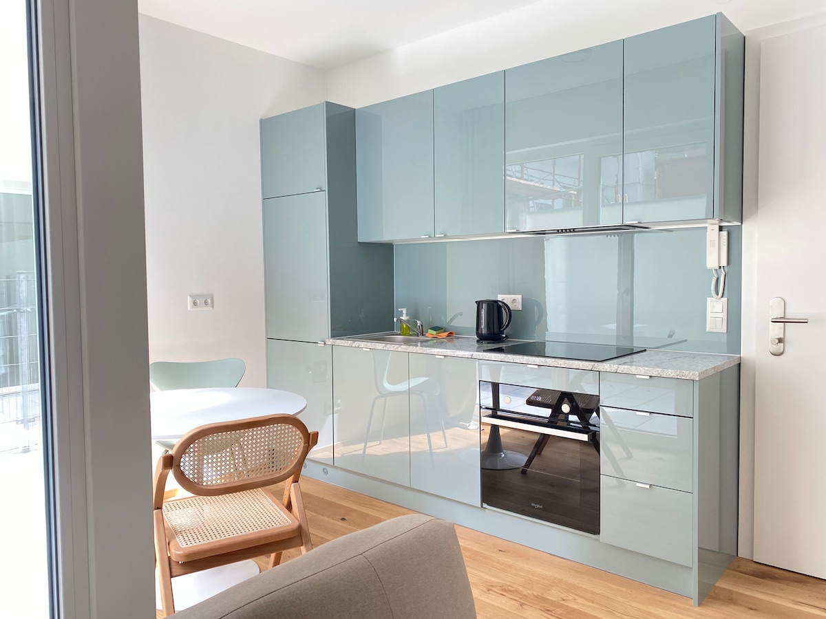 A modern kitchen area features light blue cabinets and a granite countertop. A round dining table with a pale green chair is positioned nearby. Essential appliances, including a kettle and microwave, are visible. Natural light fills the space, enhancing the minimalist design.