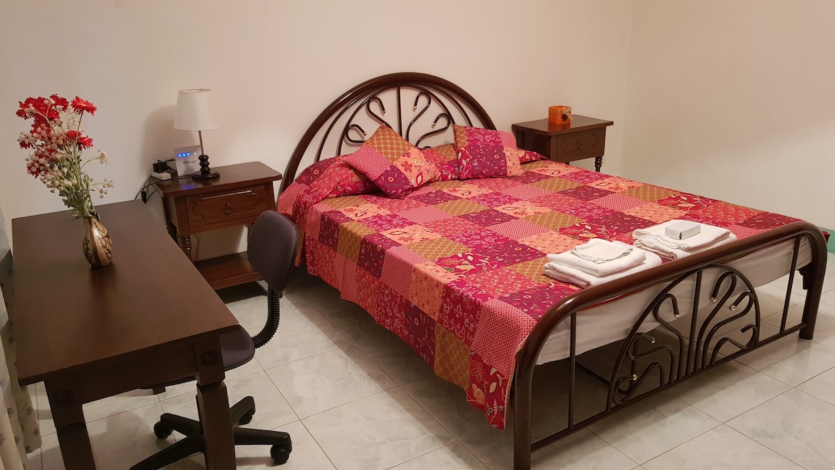 A cozy bedroom features a spacious bed adorned with a vibrant patchwork quilt and decorative pillows. A small desk and chair are positioned beside the bed, while nightstands with lamps add functionality. Fresh towels are neatly arranged at the foot of the bed.