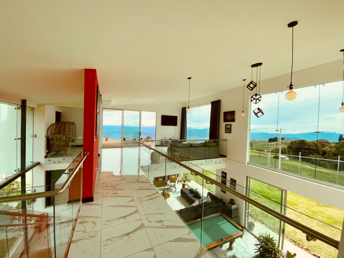 A modern interior is showcased, featuring an open space with glass walls that provide expansive views of the valley and mountains. The balcony overlooks a pool area, complemented by sleek furnishings and hanging pendant lights that illuminate the surroundings.