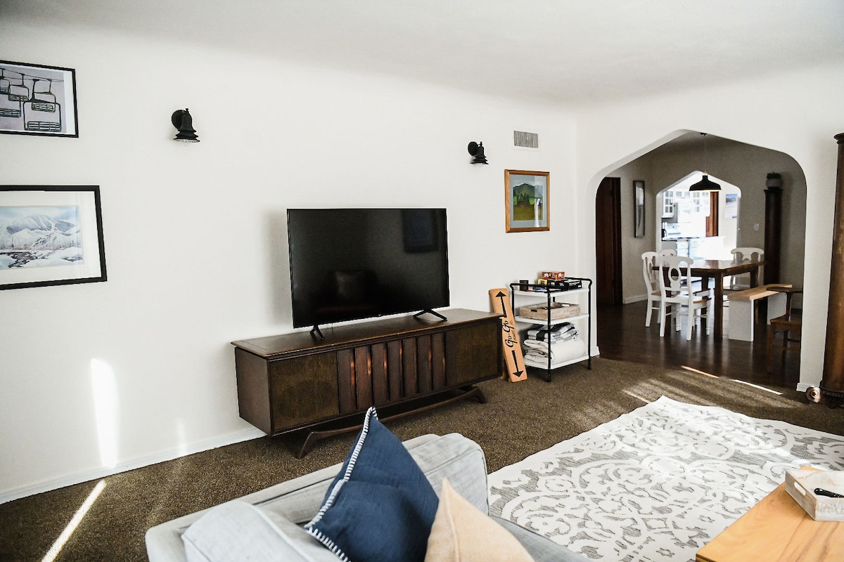 A light and airy living space features a large flat-screen television mounted above a wooden console. An inviting area rug lies on the carpeted floor, accompanied by a gray sofa adorned with decorative cushions. Arched doorways lead to a dining area visible in the background.