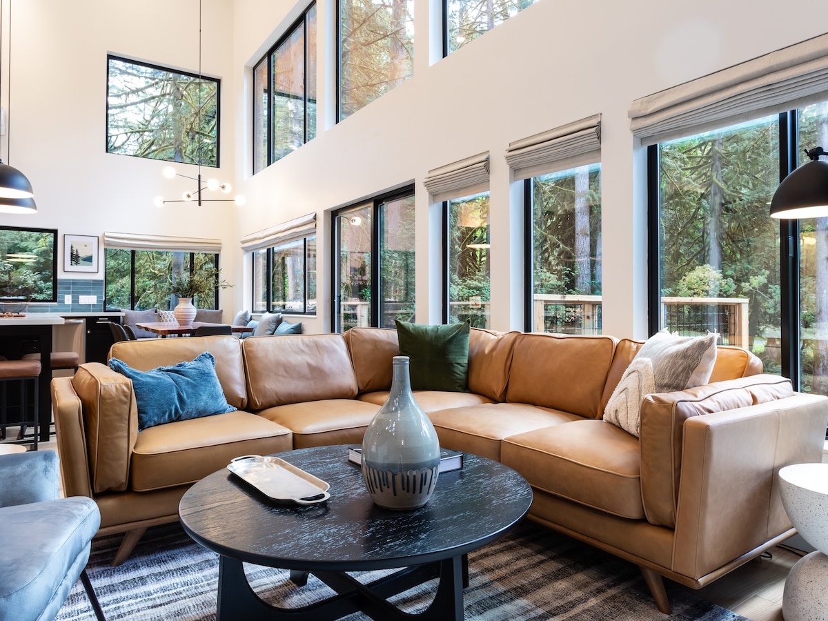 The open-concept great room features a spacious brown leather sectional sofa arranged around a circular black coffee table. Large windows provide views of the surrounding woodland, while modern lighting fixtures add an elegant touch. A cozy area rug complements the natural color palette.