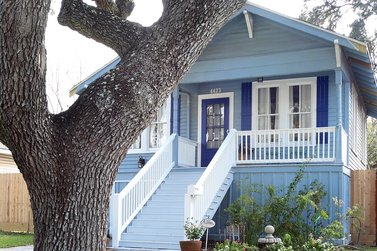 The cottage is framed by a large Live Oak tree, showcasing a light blue exterior with white railings. Stairs lead up to a welcoming entrance featuring a blue door and large windows. A garden area is visible in front, enhancing the charm of the home.