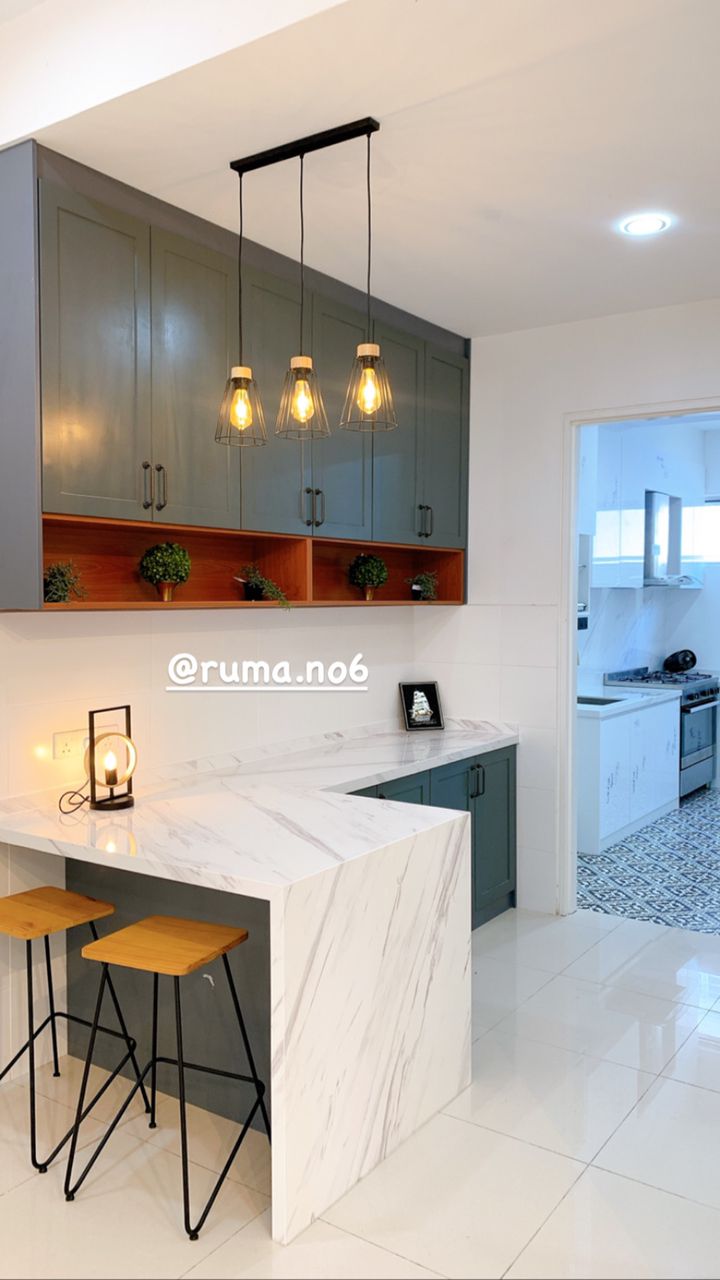 An elegant kitchen is showcased with a marble-topped bar and two wooden stools, complemented by stylish pendant lighting overhead. Upper cabinetry in muted green pairs with warm wood accents, while the backsplash features a simple yet refined design, enhancing the modern aesthetic of the space.