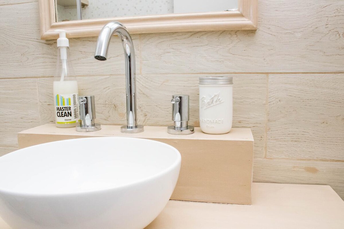 A minimalist bathroom sink area features a sleek, circular white basin positioned atop a light wooden countertop. A faucet with a modern chrome design is visible, alongside a bottle of cleaner and a jar labeled 'Soap.' Neutral-toned walls with subtle texture complement the design.