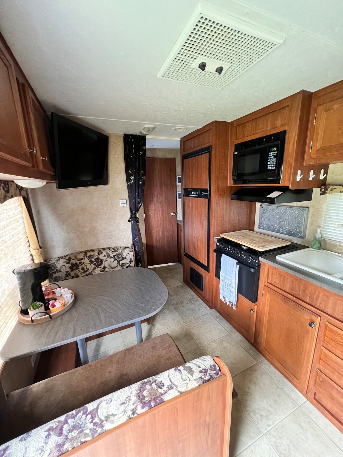 The camper's interior features wooden cabinetry and modern appliances. A compact table with cushioned seating is positioned near a window. A microwave and stove are integrated into the kitchen area, which also includes a sink and cabinetry for storage.