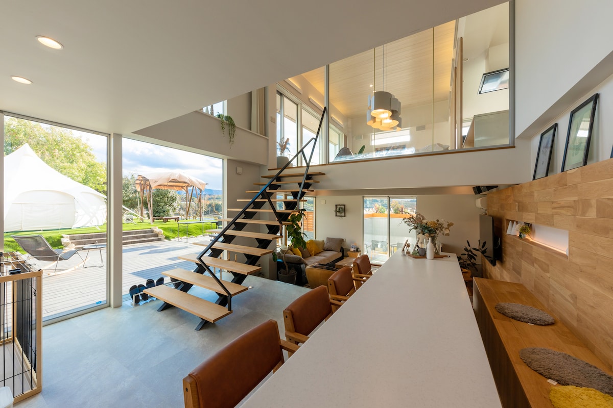 An open-concept living and dining area is showcased with large glass windows, allowing natural light. A modern staircase with wooden treads leads to the upper level. The space features minimalist decor, a cozy seating arrangement, and views of the outdoor patio and tent area.