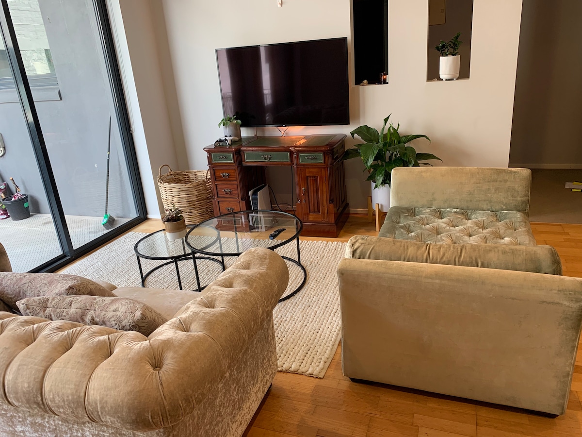 A cozy living area features two plush armchairs and a tufted couch, arranged around a glass coffee table. A television is mounted on the wall opposite, with a wooden cabinet situated beside it. Natural light fills the space through sliding glass doors, enhancing the warm tones.