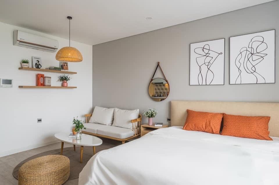 A bright bedroom features a spacious bed with orange accent pillows. A cozy seating area includes a light-colored sofa and a round coffee table. An air conditioning unit is mounted on the wall, and two minimalist art pieces are displayed above the bed.