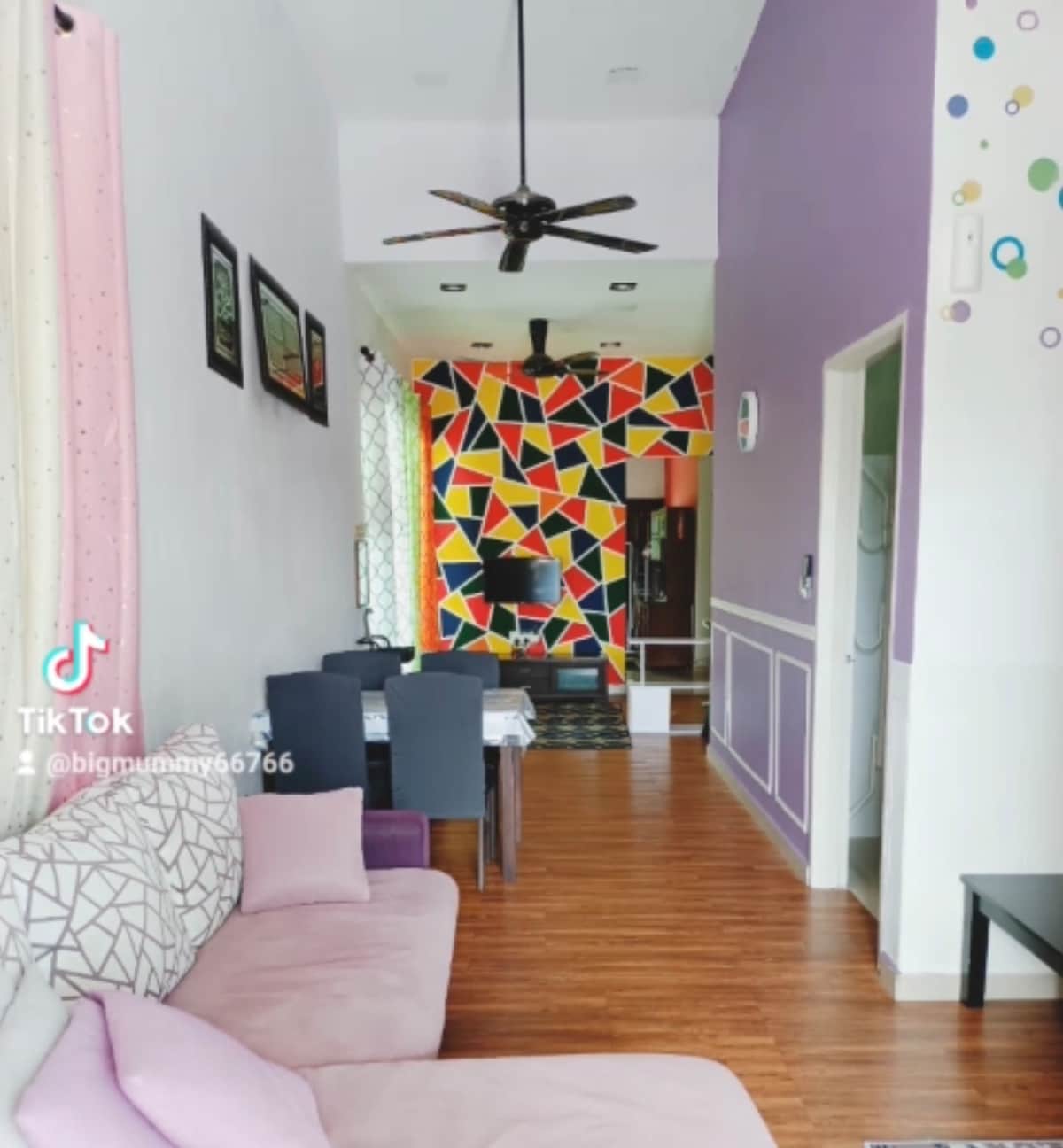 A comfortable living space features light-colored walls and a bold geometric accent wall. A spacious seating area is complemented by a dining table and dark chairs. Wooden flooring contributes to the overall warmth of the environment, while a ceiling fan is visible for added comfort.