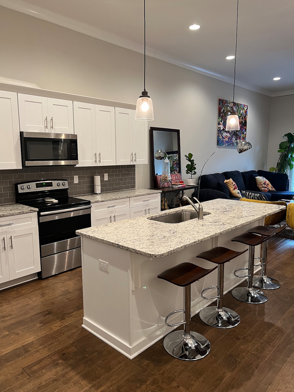 A modern kitchen features sleek white cabinetry and stainless steel appliances, with a spacious granite countertop providing ample prep space. Four contemporary bar stools are positioned at the counter. Soft lighting hangs above the area, creating an inviting ambience.