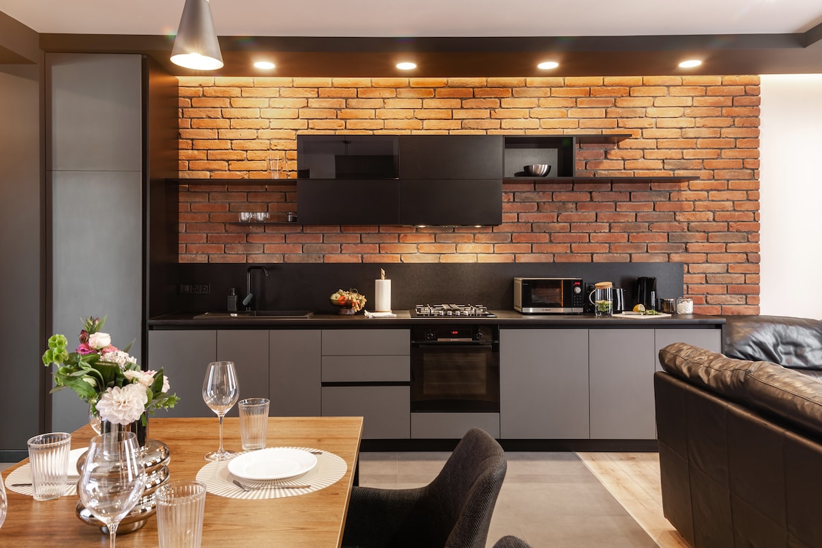 A modern kitchen area features dark cabinetry harmoniously paired with a warm brick accent wall. A dining table is set nearby, adorned with elegant glassware and a simple floral centerpiece. Appliances including a microwave and kettle are visible on the countertop.