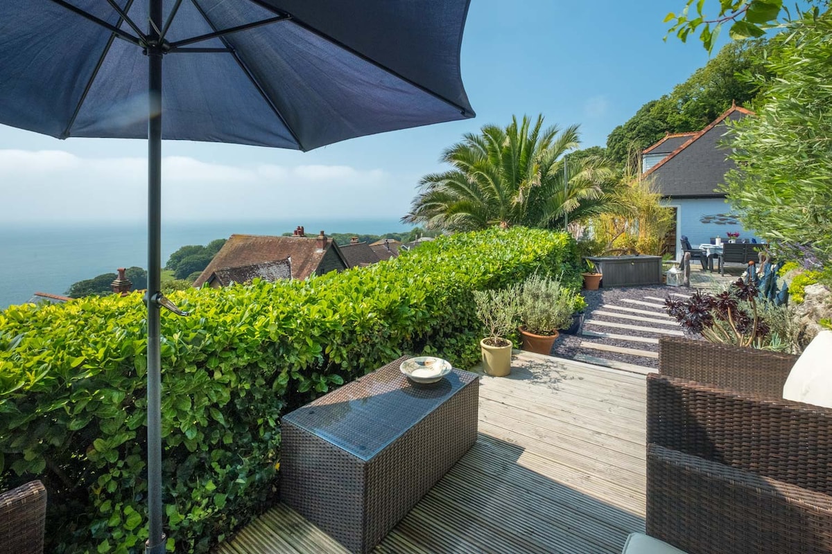 An outdoor terrace is shown, featuring a wicker seating area and a circular table with chairs. A large umbrella provides shade while vibrant green foliage frames the scene. The sea is visible in the distance, complemented by a clear blue sky.