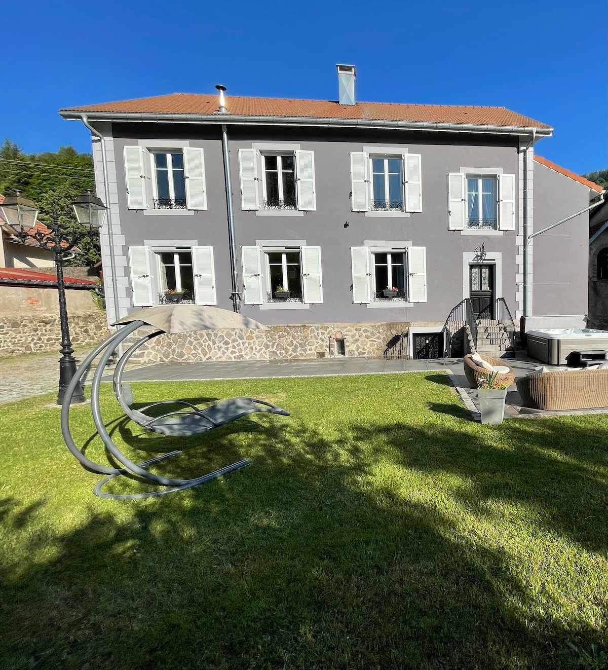 The two-story gray house, set against a clear blue sky, features white shutters and four windows with flowers. A manicured lawn surrounds the home, while a cozy outdoor seating area and a jacuzzi are visible, providing a relaxing space in the garden.