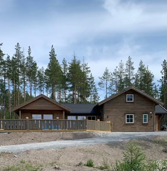 A wooden cabin is nestled among tall trees, featuring a spacious deck at the front. The structure includes a mix of gabled roofs and large windows, allowing natural light to complement the earthy tones of the facade.