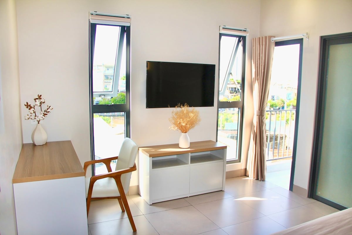 A modern room features large windows that allow natural light to fill the space. A flat-screen TV is mounted on the wall opposite a wooden desk, accompanied by a white chair. A decorative vase with dried flowers sits on the desk, adding a touch of elegance.