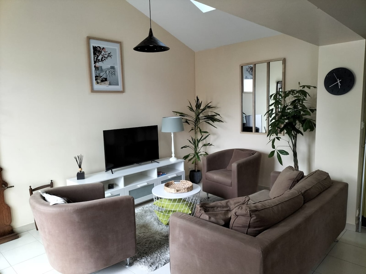 The living area features a comfortable arrangement of two armchairs and a spacious sofa, complemented by a central coffee table. A television sits on a white console, while potted plants and a mirror add natural elements to the warm ambiance. Soft light enters through the skylight.